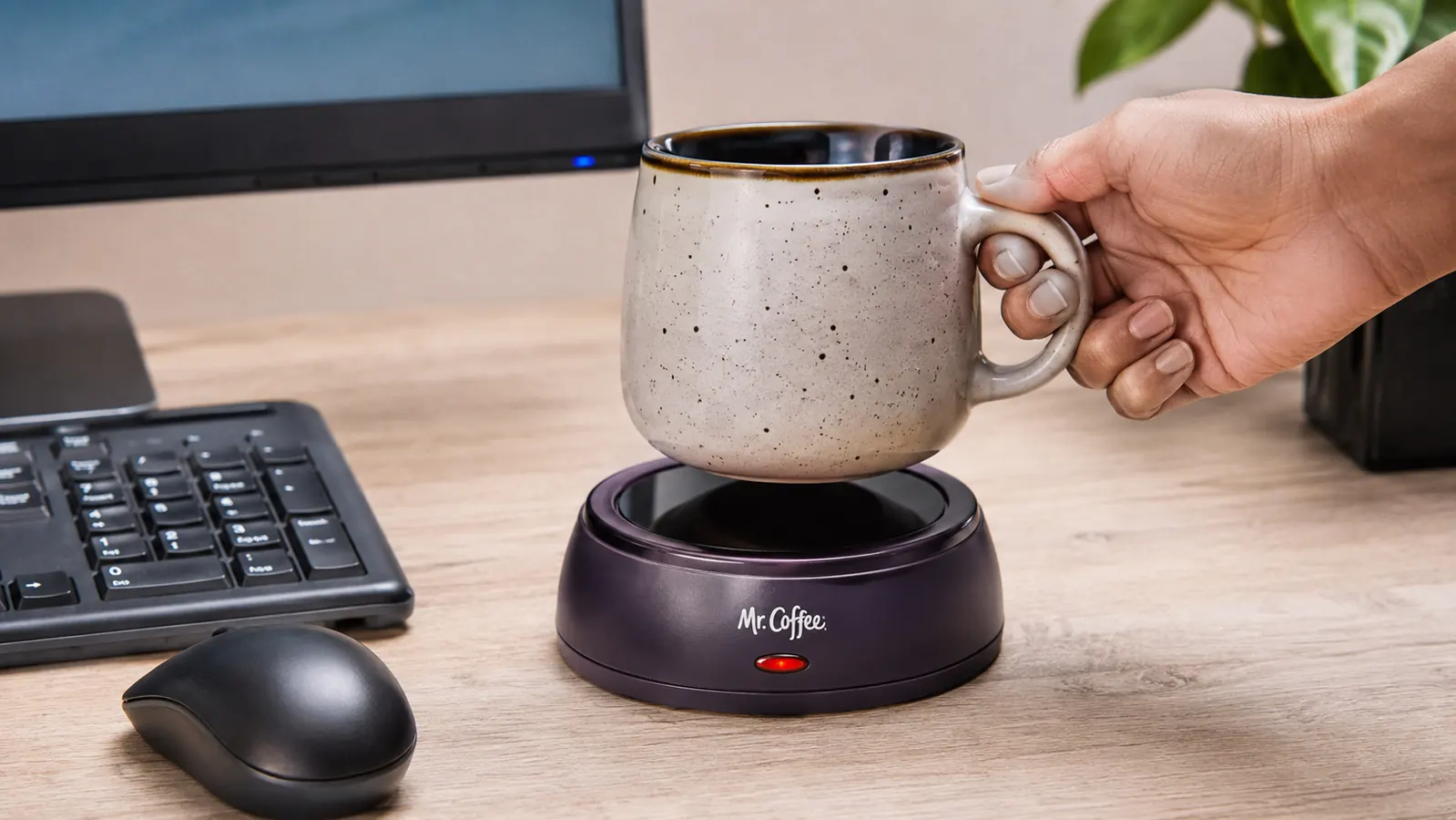 A hand setting a ceramic mug down onto the Mr. Coffee mug warmer plate next to a keyboard