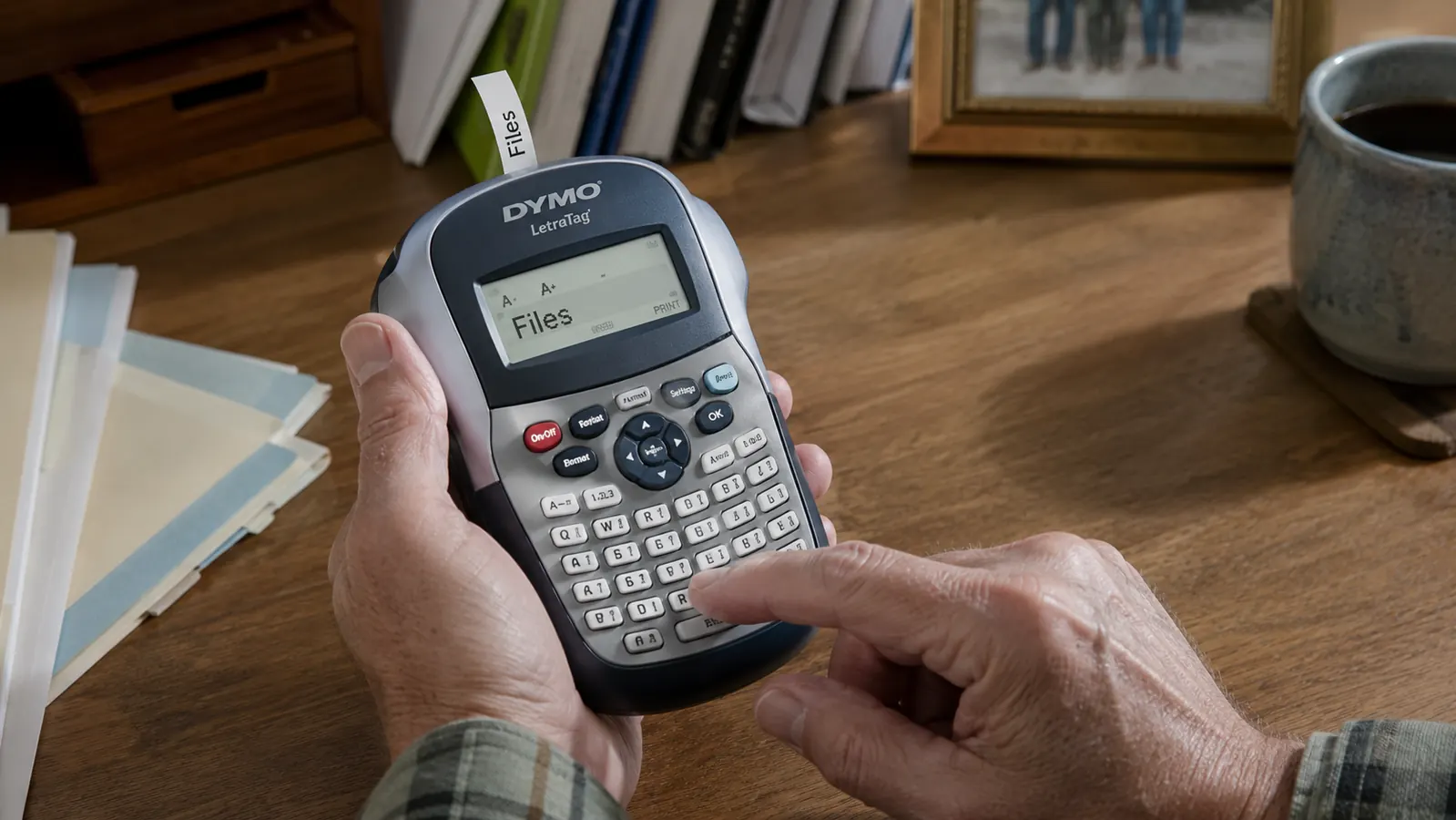 A hand printing a label with a handheld DYMO label maker at a home office desk